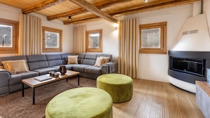 Un salon moderne avec un canapé d’angle gris, une table basse en bois, deux poufs verts, une cheminée et de grandes fenêtres avec des rideaux.