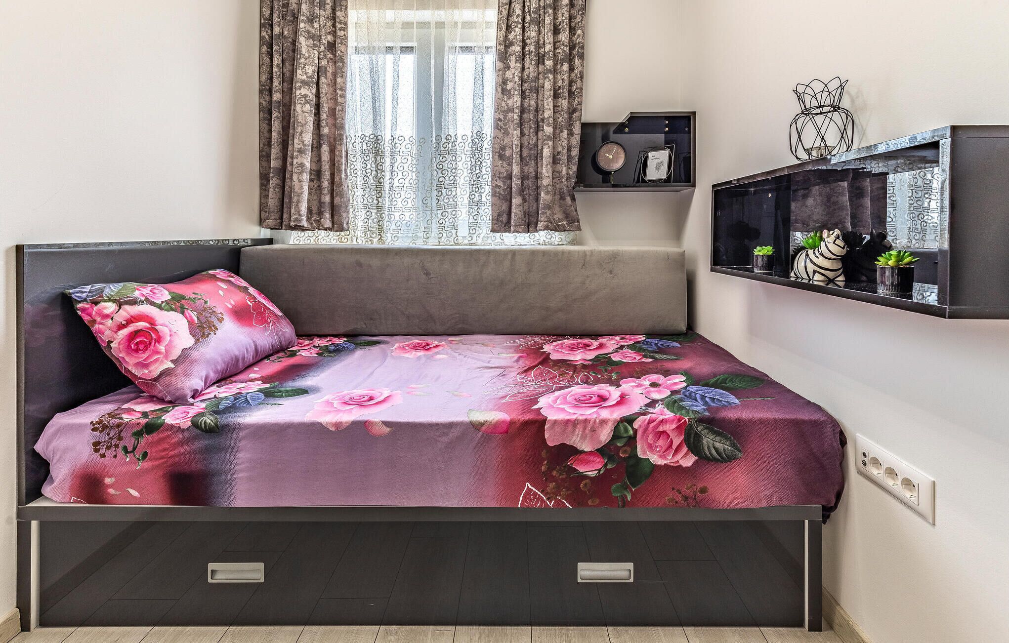 Une chambre à coucher avec un lit, une fenêtre avec des rideaux, une table de chevet avec une horloge et une étagère avec des objets décoratifs.