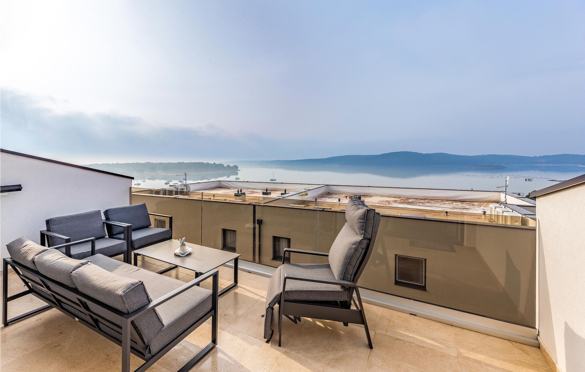 A rooftop terrace with a sofa, chair, and table, overlooking a calm body of water and distant hills.