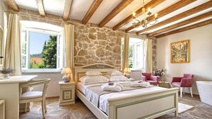 Una camera da letto con parete in pietra, travi in legno, un letto con lenzuola bianche, una scrivania, sedie e un quadro.