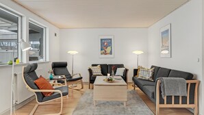 Een moderne woonkamer met een bank, fauteuils, een salontafel en vloerlampen. Er hangen posters aan de muur en een raam met uitzicht.