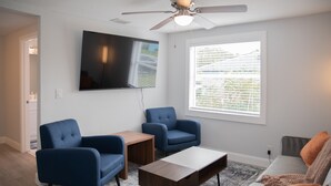 A modern living room with a flat-screen TV, two blue armchairs, a wooden coffee table, and a grey sofa with orange cushions.