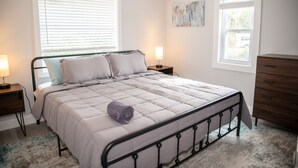 A bedroom with a bed, two bedside tables, a dresser, and two windows with blinds.