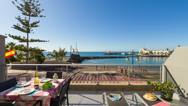 A terrace with a table set for dining, overlooking a coastal area with a clear view of the sea and a pier.