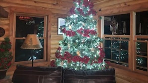 Interior acogedor de una cabaña con techo de madera, un árbol de Navidad decorado con luces y flores rojas, una lámpara y vistas del cielo nocturno a través de amplios ventanales.