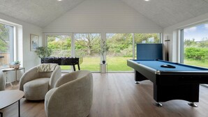 Ein Zimmer mit Poolbillard, zwei Stühlen, einem Kicker und großen Fenstern mit Blick ins Grüne.