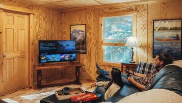 A cozy room with wooden walls, a television, a Scrabble game, and a person relaxing on a couch.