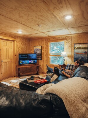 Ein gemütliches Zimmer mit Holzwänden, einem Fernseher, einem Scrabble-Spiel und einer Person, die auf einem Sofa entspannt.