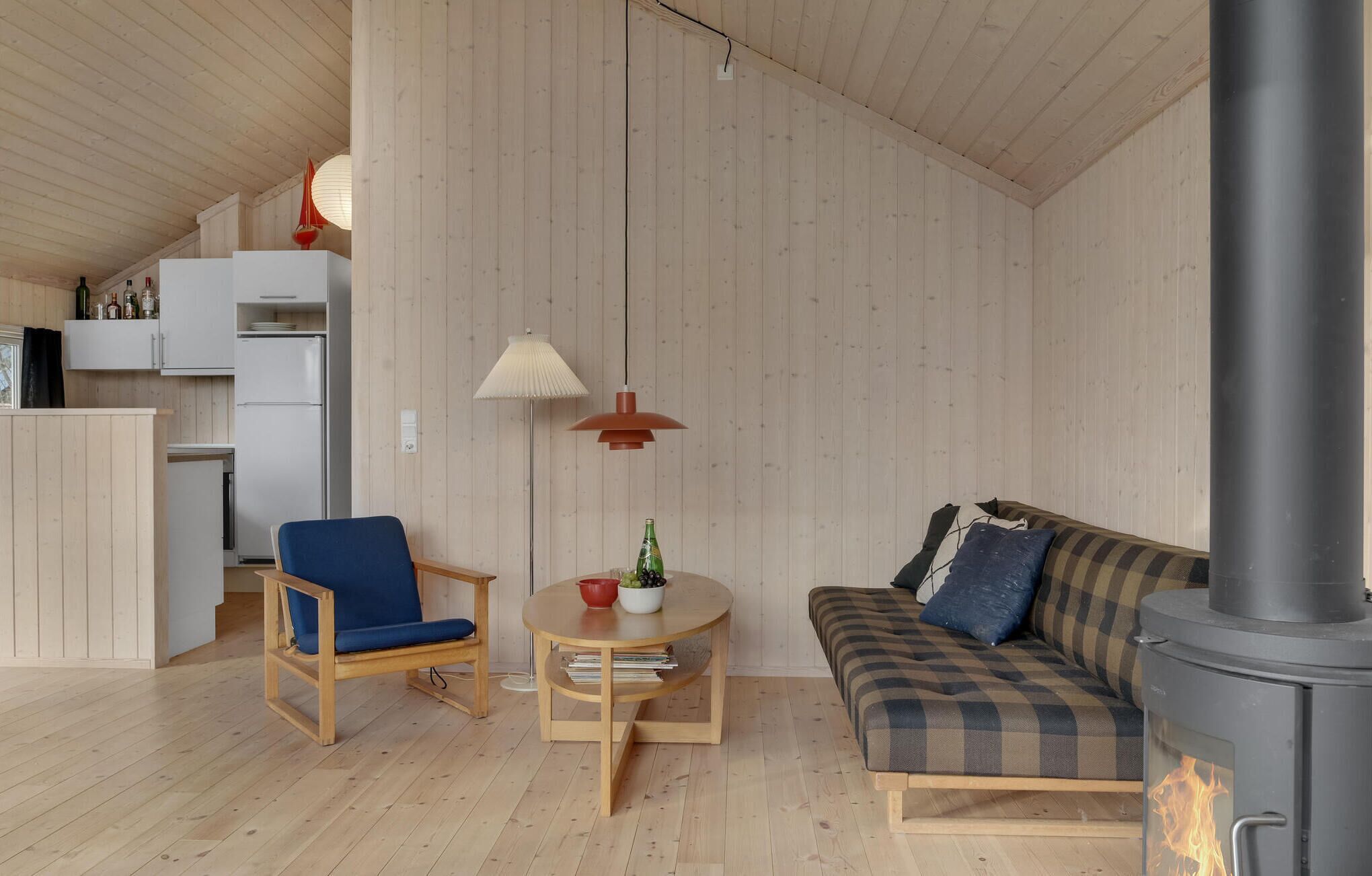 Una habitación acogedora con piso de madera, una silla azul, una mesa redonda de madera con una lámpara, un sofá a cuadros y una estufa de leña a la vista.