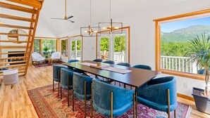 A spacious dining area with a long table, blue chairs, and a view of lush greenery through large windows. - Luxury Home with MountainViews•PoolTable•Chefs Kitchen•FirePit (Lake Lure)