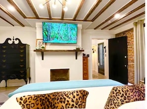 A bedroom with a bed, a TV mounted above a fireplace, a wardrobe, and a door leading to another room.