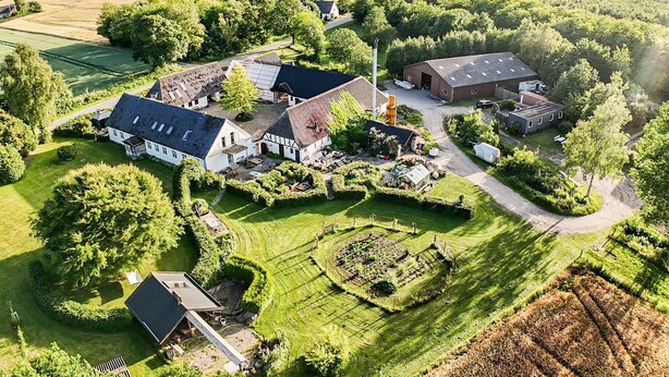 Luchtbeeld van een landelijk gelegen bezit met meerdere gebouwen, waaronder een groot huis met een schuin dak, kleinere constructies en een omheind tuingedeelte.
