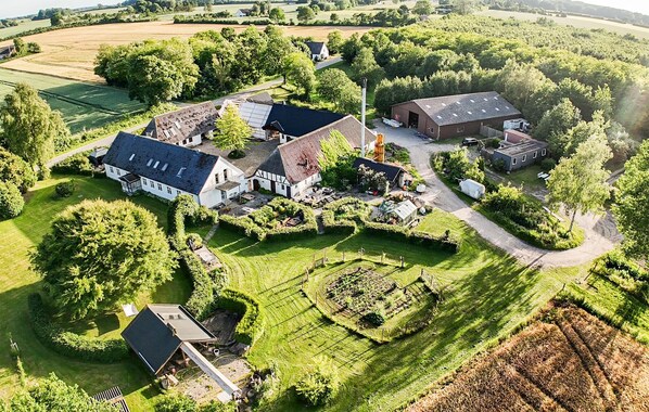 An aerial view of a rural property with multiple buildings, including a large house with a sloped roof, smaller structures, and a fenced garden area.