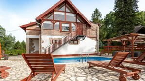 Una cabaña de dos pisos con un patio de piedra, una piscina y sillas de madera.