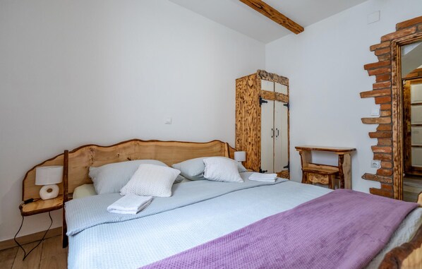 Un dormitorio con cabecera de madera, cama con sábanas blancas, mesita de noche de madera con lámpara, una pequeña mesa de madera y un armario con espejo.
