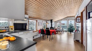 A modern kitchen with a wooden ceiling, large windows, and a dining area with chairs and a table.