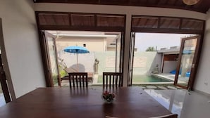 Una mesa de comedor con cuatro sillas, un florero con flores y vistas a una piscina y una zona de estar exterior a través de puertas de cristal.
