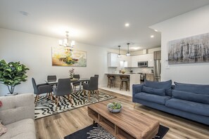 Un salon moderne avec un canapé bleu, un coin repas agrémenté d’un tapis à motifs zébrés, et une cuisine dotée de meubles blancs et d’appareils électroménagers en acier inoxydable.