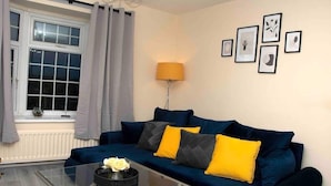 Una sala de estar con un sofá azul oscuro, cojines amarillos, una mesa de centro de cristal con un jarrón de flores, una lámpara y cuadros en la pared.