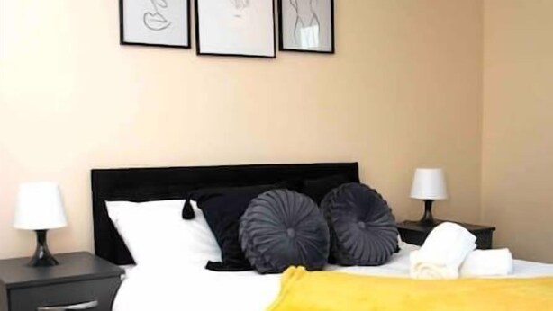 Un dormitorio con cabecera negra, ropa de cama blanca, una manta amarilla y tres cuadros en la pared.