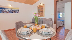 Zona de menjador amb taula rodona preparada per l'esmorzar, amb plats, verres de suc d'aranja i pastisseria. Hi ha un ram de flors roses sobre la taula i un quadre de paisatge a la paret.