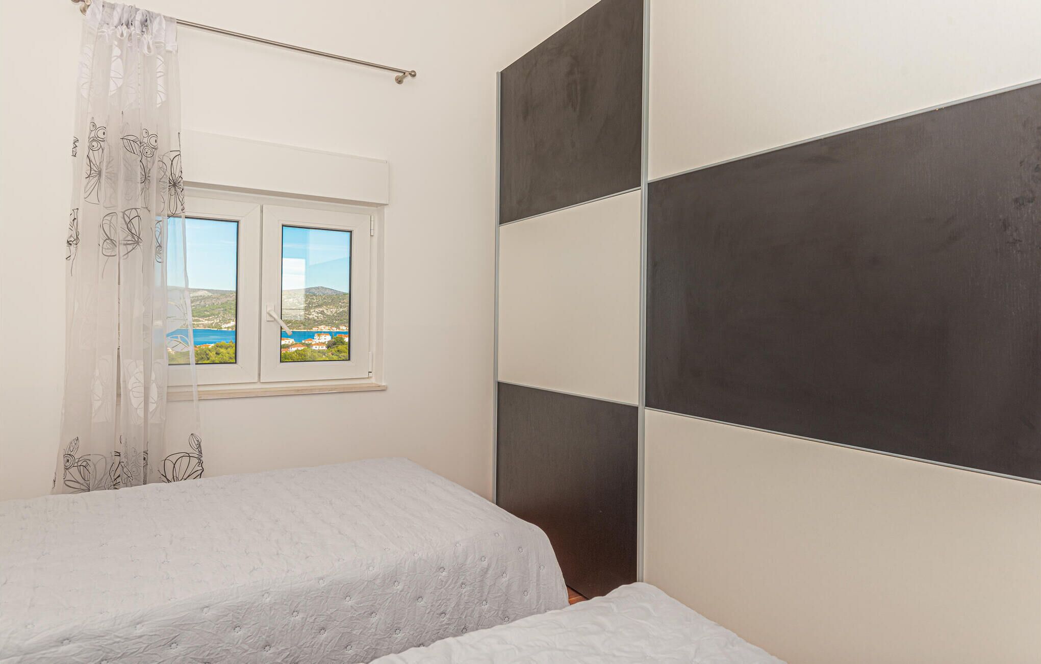 Un dormitorio con dos camas, una ventana con vistas al paisaje costero y un muro con paneles oscuros grandes.