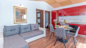 Un saló modern amb un sofà gris, una taula de dinar amb cadires i una cuina amb armaris vermells.