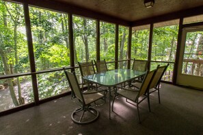 Una mesa con cubierta de vidrio y sillas de metal en una habitación con ventanas amplias que dan a un bosque.