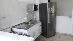 A kitchen with white cabinets, a stainless steel refrigerator, a microwave, and a sink with a yellow sponge. - 500m da Praia, BIWA Edifício com Piscina, 1garagem, WI-FI, Portaria 24hs, churrasqueira, playground, jardim, Centro do Guarujá, SP (Guarujá)