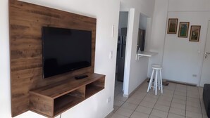 A modern living room with a large flat-screen TV mounted on a wooden wall unit, a white stool, and a door leading to another room. - 500m da Praia, BIWA Edifício com Piscina, 1garagem, WI-FI, Portaria 24hs, churrasqueira, playground, jardim, Centro do Guarujá, SP (Guarujá)