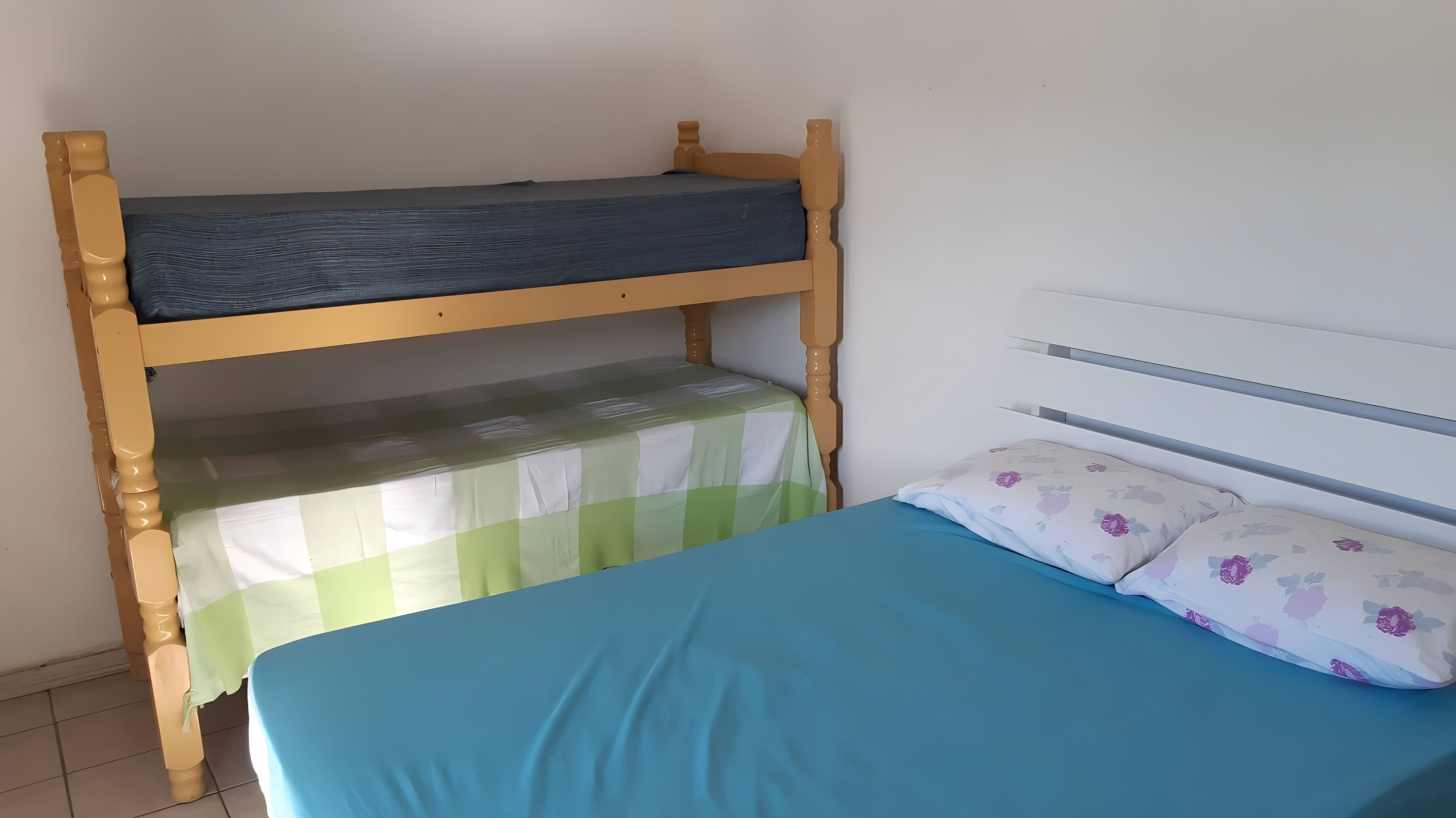 A bunk bed with a green checkered bedspread and a blue mattress, a white headboard, and a floral pillow.