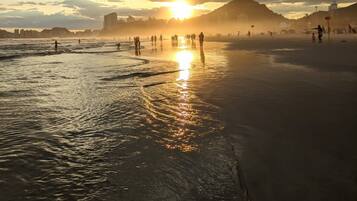 A beach scene with the sun setting over the horizon, people walking along the shore, and a distant city skyline.
