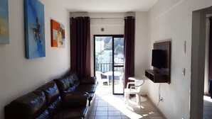 A modern living room with a leather sofa, a flat-screen TV mounted on the wall, and a balcony with a view of the city. - 500m da Praia, BIWA Edifício com Piscina, 1garagem, WI-FI, Portaria 24hs, churrasqueira, playground, jardim, Centro do Guarujá, SP (Guarujá)