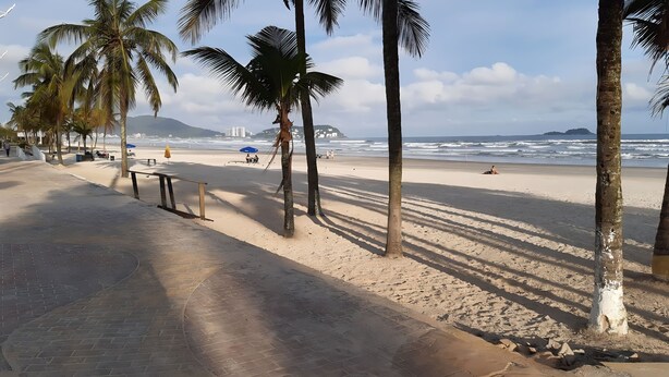 A beach with palm trees, a paved walkway, and the ocean in the background. - 500m da Praia, BIWA Edifício com Piscina, 1garagem, WI-FI, Portaria 24hs, churrasqueira, playground, jardim, Centro do Guarujá, SP (Guarujá)