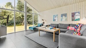 Een moderne woonkamer met een grote glazen deur die uitkomt op een tuin, een grijze bank, een salontafel en kunstwerken aan de muur.