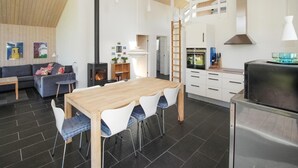 Een moderne keuken met een centraal eiland, een eetgedeelte met witte stoelen, een slaapverdieping met een houten trap, en een woonkamer met een bank en een open haard.