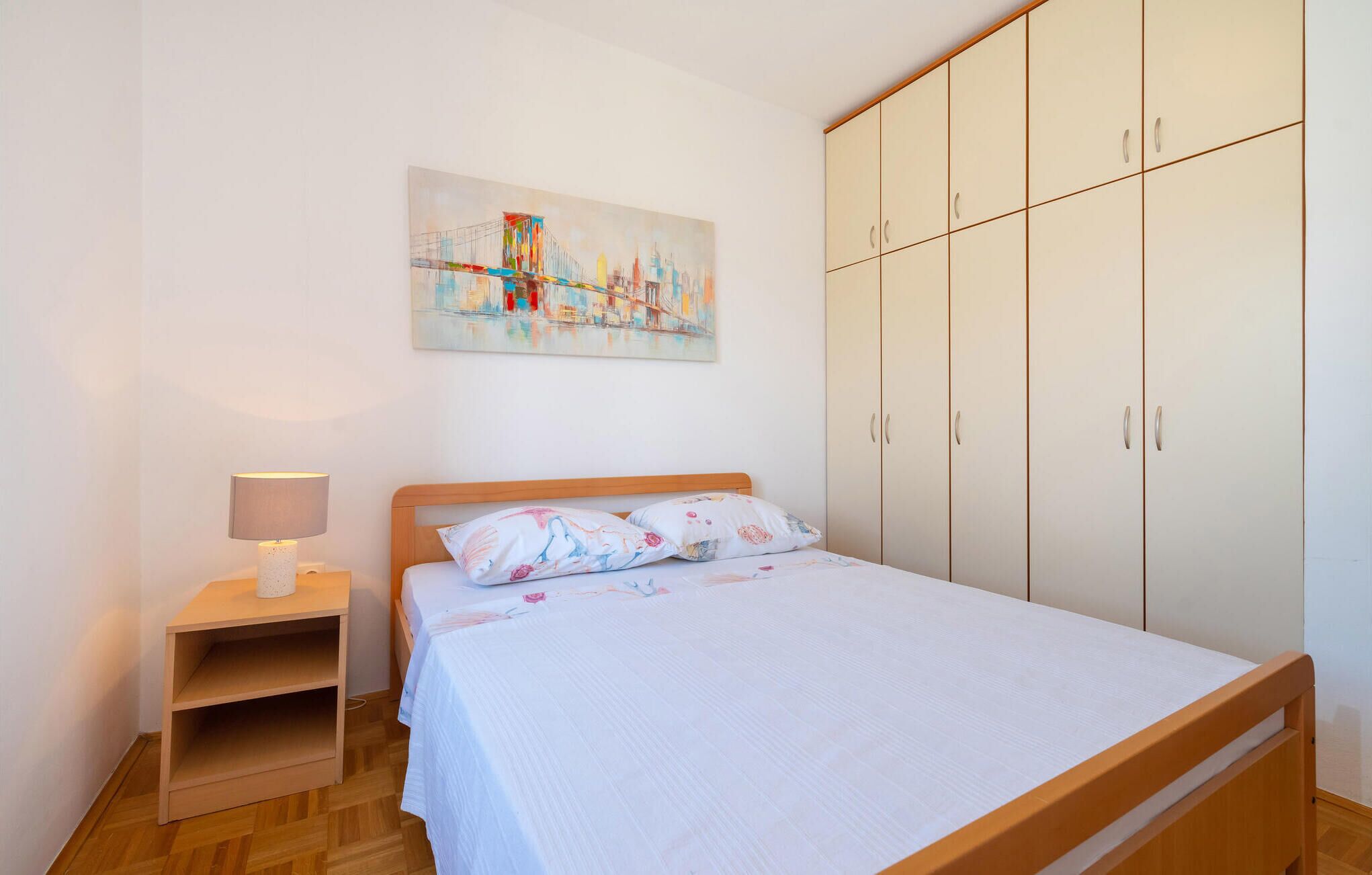 Un dormitorio con cama, mesita de noche, lámpara, armario y un cuadro de paisaje urbano en la pared.