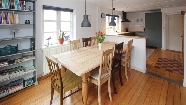 A dining area with a wooden table and chairs, a bookshelf with books, a window with a view, and a kitchen area with a sink and stove. - Cozy home in Samsø with WiFi (Samsø)