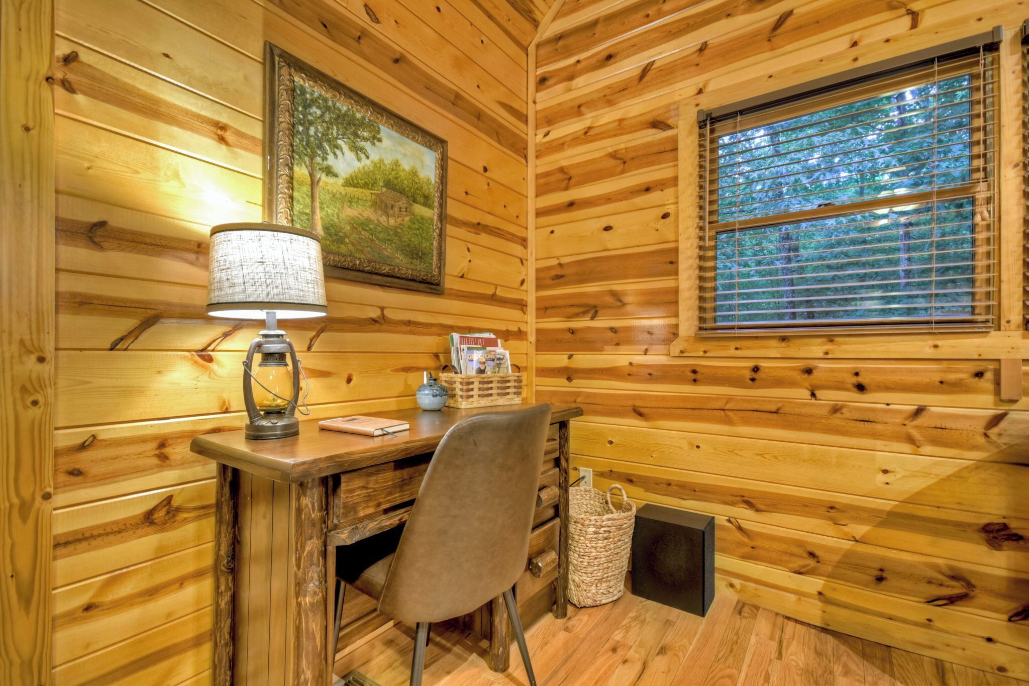 L’intérieur d’un chalet en bois, avec un bureau, une chaise, une lampe, un tableau encadré, une fenêtre et un panier.