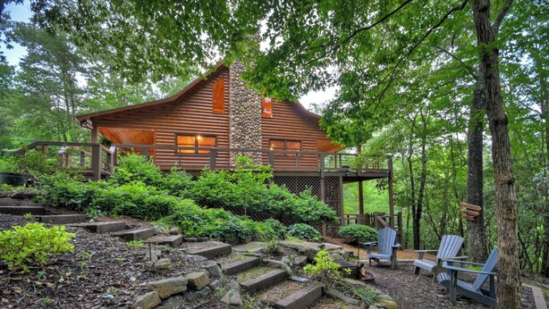 A log cabin with a stone chimney, surrounded by a garden with steps leading up to the house, and two Adirondack chairs near the entrance.