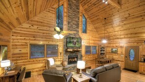 A log cabin interior with a fireplace, wooden walls, a ceiling fan, and a dining area.