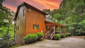 A wooden log cabin with a porch and stairs leading to a door, surrounded by lush greenery.