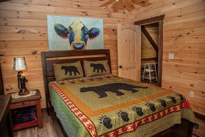 Une chambre à coucher dans une cabane en bois, avec un lit recouvert d’une courtepointe à motifs d’ours, un tableau représentant une vache et une petite table avec une lampe.
