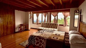A bedroom with a wooden ceiling, a large bed with patterned bedding, a nightstand, and a view of greenery outside. - Posada Quetzalin (Cuetzalan del Progreso)