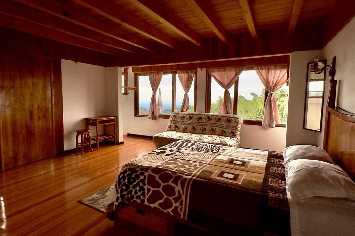A bedroom with a wooden ceiling, a large bed with patterned bedding, a nightstand, and a view of greenery outside.