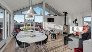 A spacious living area with a large round table, modern furniture, a fireplace, and large windows offering a view of the outside. - Amazing home in Løkken with kitchen (Løkken)