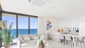Un salon moderne doté d’une grande fenêtre donnant sur l’océan, une cuisine avec des meubles blancs et un coin repas comprenant une table et des chaises blanches.