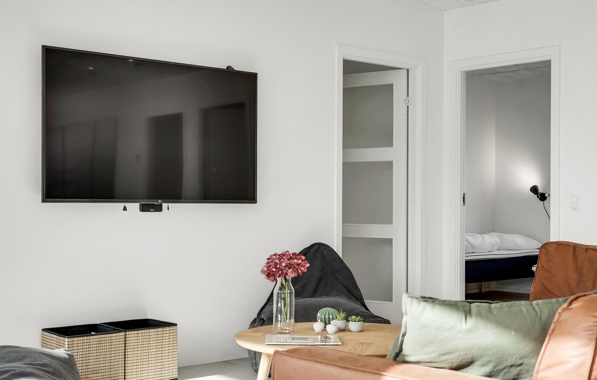 A modern living room with a flat-screen TV mounted on the wall, a small table with a vase of flowers, and a bed in the adjacent room.