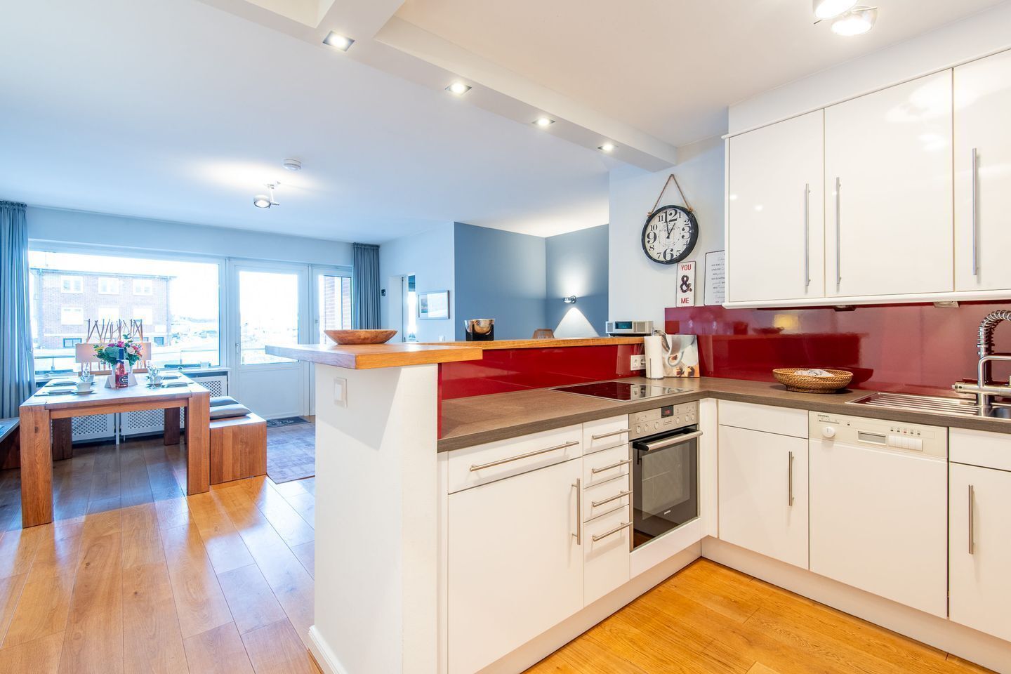 A modern kitchen with white cabinets, a red countertop, and a built-in oven. There is a dining area with a wooden table and chairs, and a large window providing natural light.