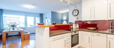 A modern kitchen with white cabinets, a red countertop, and a built-in oven. There is a dining area with a wooden table and chairs, and a large window providing natural light.